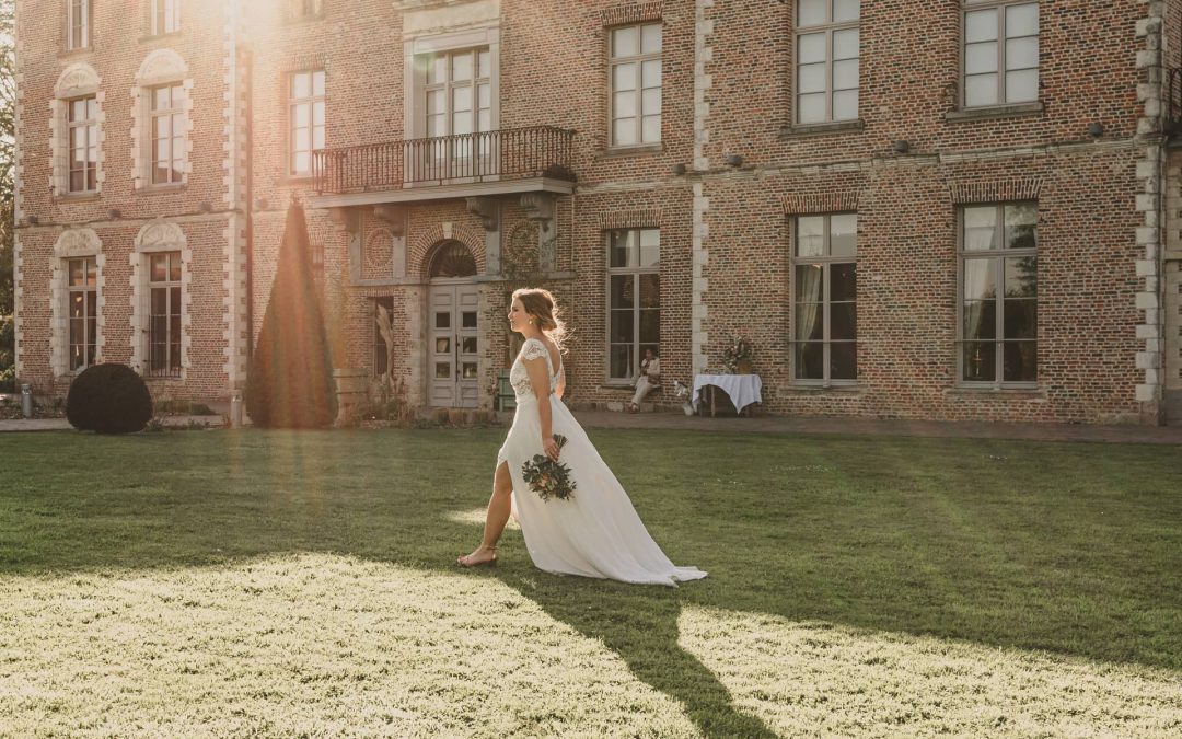 Une personne en robe de mariée blanche tenant un bouquet marche sur une pelouse devant un bâtiment en briques.