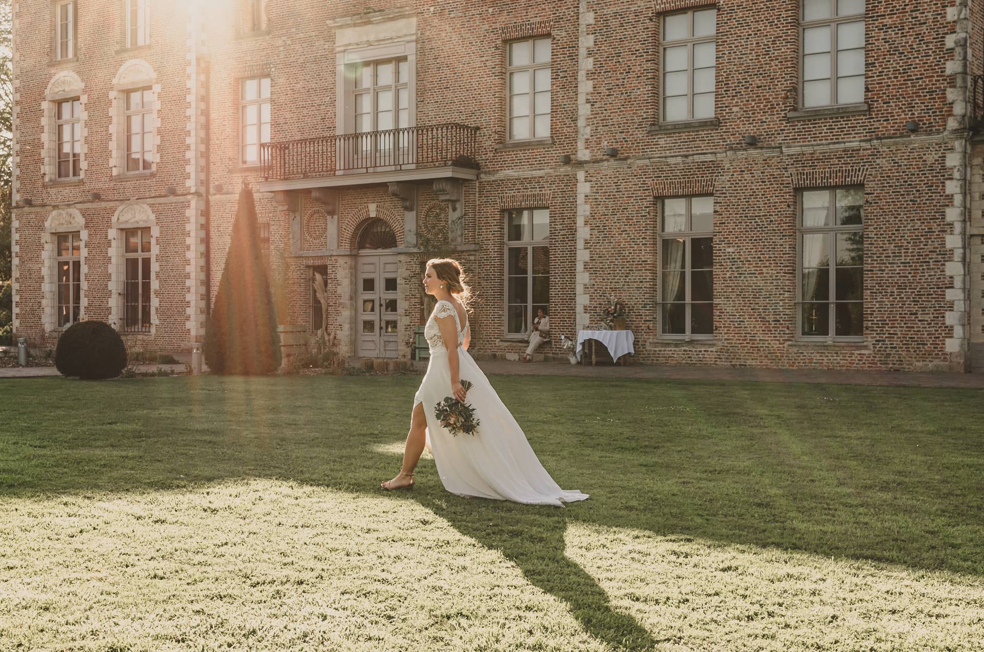 Une personne en robe de mariée blanche tenant un bouquet marche sur une pelouse devant un bâtiment en briques.