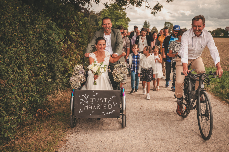La mariée est poussée dans un chariot vélo avec l’inscription “JUST MARRIED”, une autre personne à vélo à côté d’elle, entourée de plusieurs personnes célébrant l’occasion.