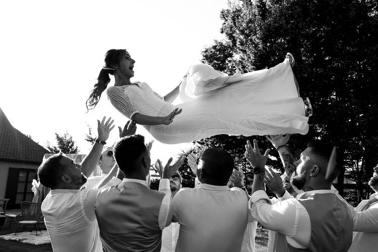 Une mariée en robe fluide est soulevée dans les airs par plusieurs invités lors d’un mariage en plein air.