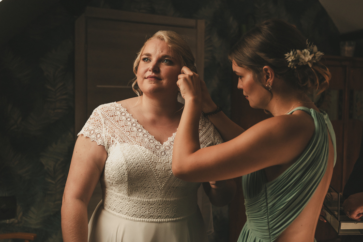 Une mariée en robe blanche avec des détails en dentelle, aidée par une autre personne en robe verte.