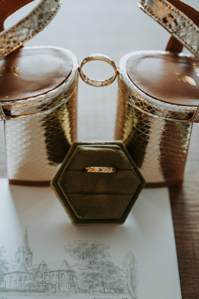 Détails de mariage de Madeline et Julien : alliances présentées sur des chaussures dorées et un écrin en velours vert.