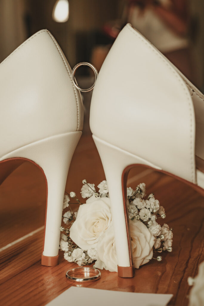 Détails de mariage : alliances de Manon et Firmin balancées entre deux escarpins blancs avec des roses.