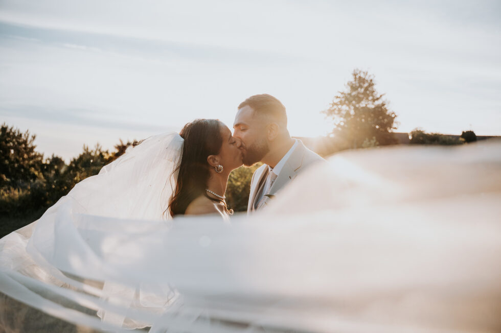 Baiser passionné des mariés avec un effet de voile blanc volant au premier plan créant un flou artistique.