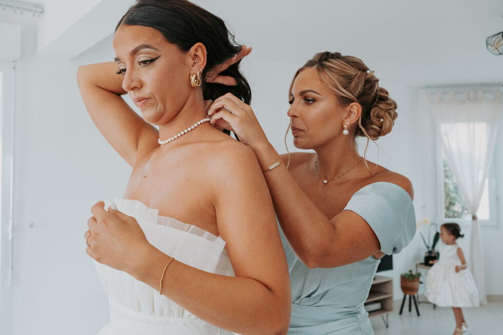 Une demoiselle d'honneur aide la mariée à attacher son collier de perles blanches.