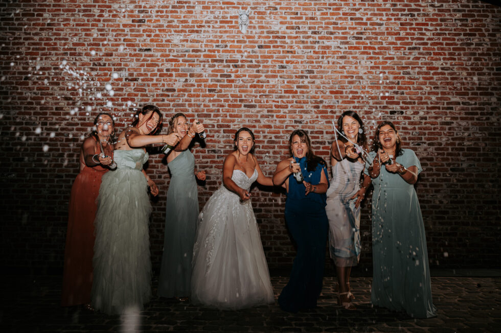 La mariée et ses amies s'amusent à arroser la soirée avec du champagne (Champagne Shower) devant un mur de briques.