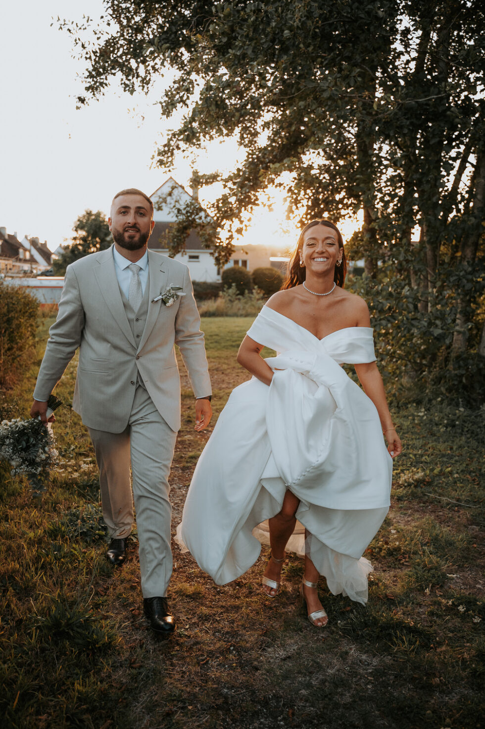 Les mariés marchent souriants dans l'herbe. La mariée porte sa seconde tenue, une robe longue en satin à épaules dénudées.