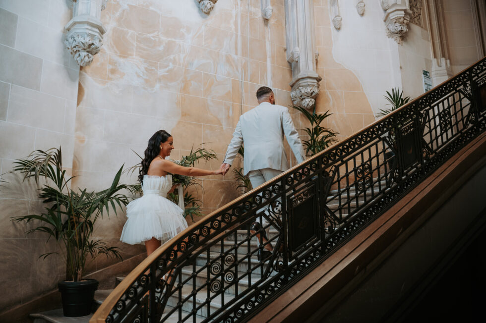 Le marié et la mariée (en robe courte) montent main dans la main un grand escalier d'honneur en pierre.