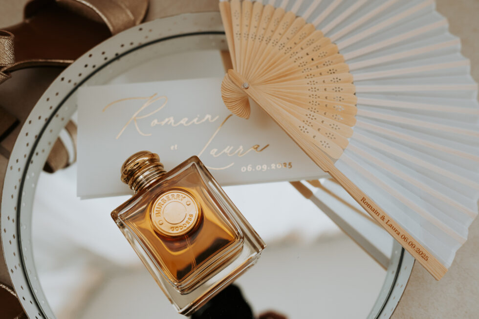 Composition de détails mariage : Parfum Burberry, éventail blanc et faire-part au nom de Romain et Laura.