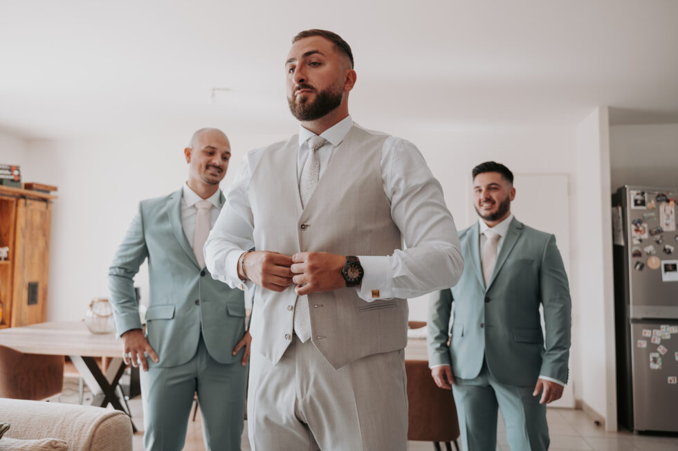 Le marié en costume beige boutonne son gilet, entouré de ses deux témoins en costumes vert d'eau.