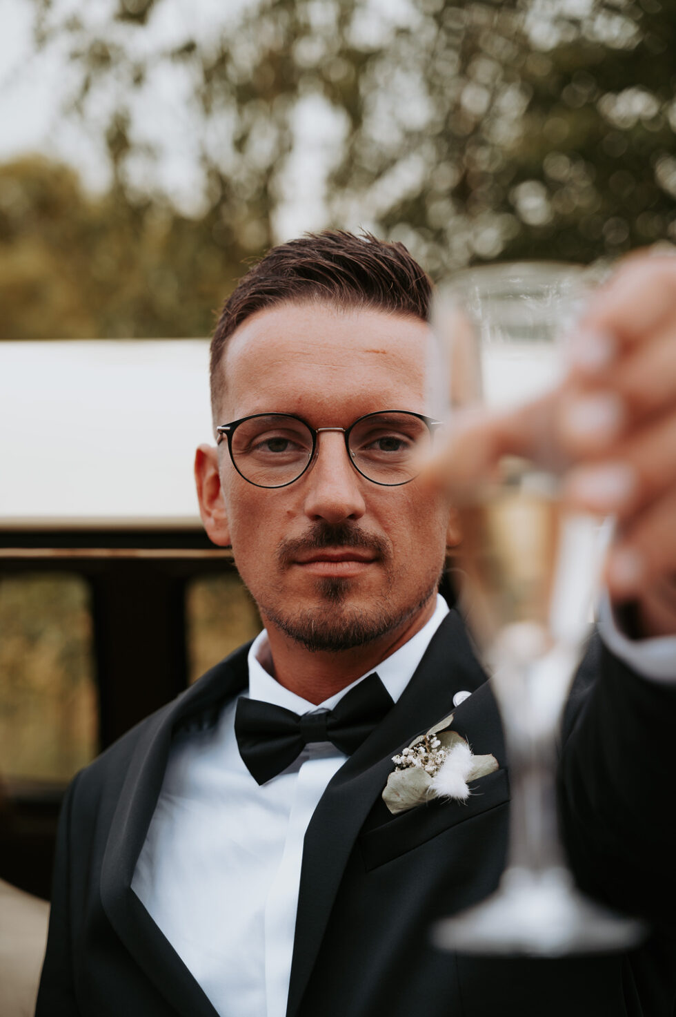 Portrait du marié avec lunettes, levant sa coupe de champagne vers l'objectif (flou au premier plan).