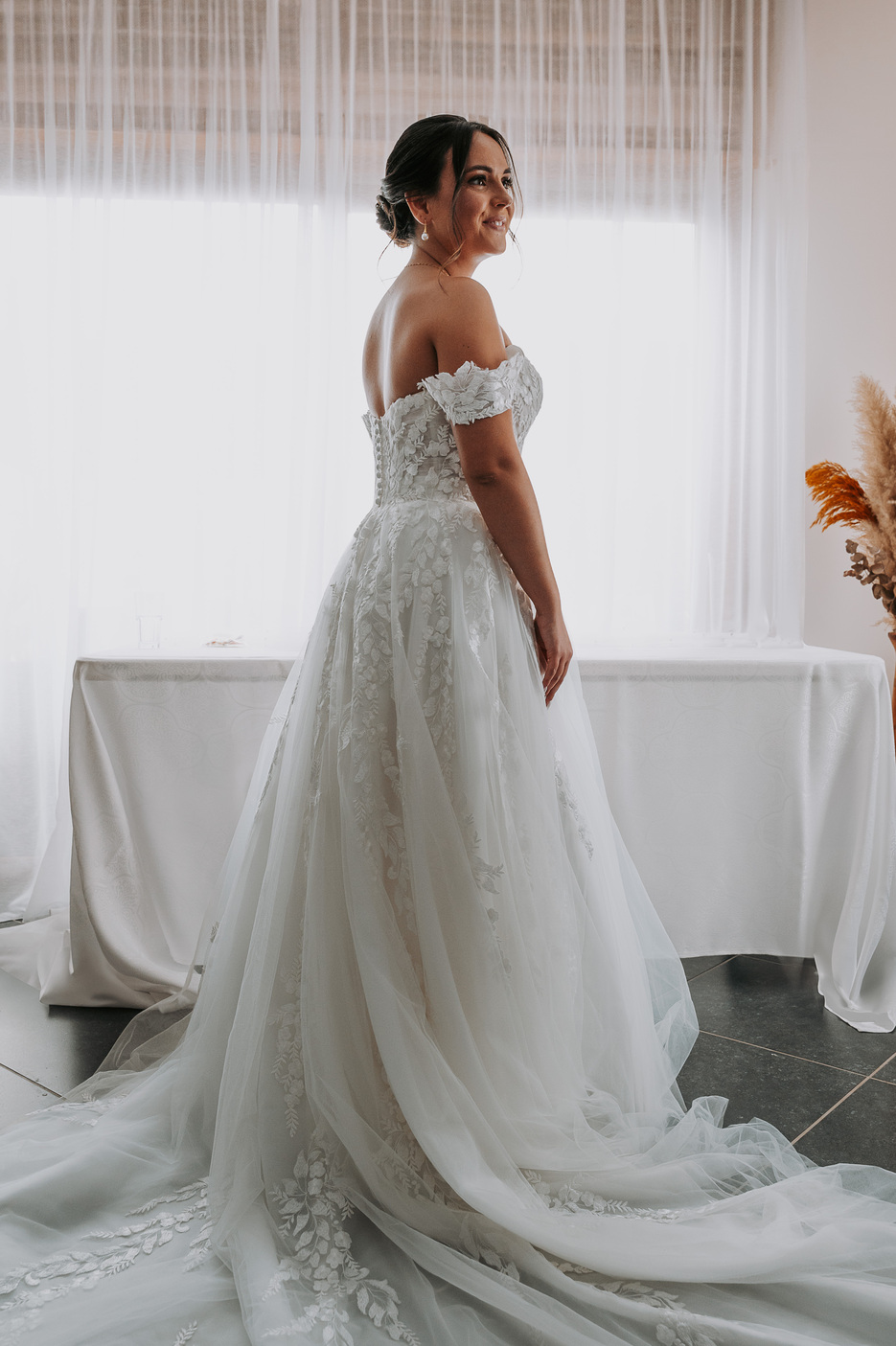 La mariée vue de dos, présentant les détails de sa robe en dentelle à épaules dénudées et sa traîne.