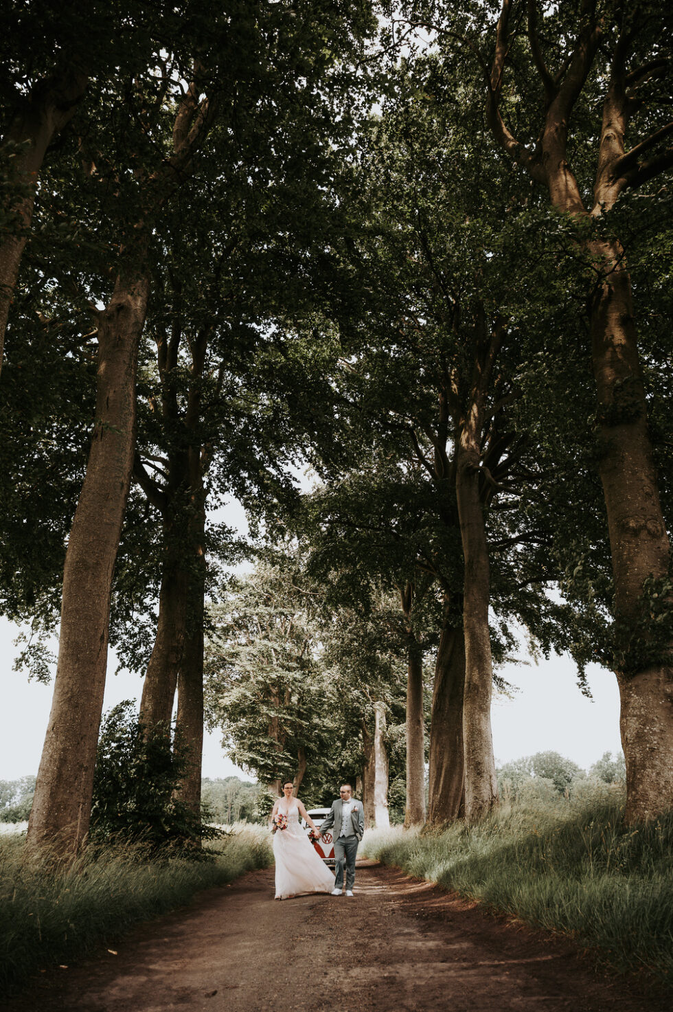 Les mariés marchent main dans la main dans une allée forestière avec un Combi Volkswagen vintage en arrière-plan.