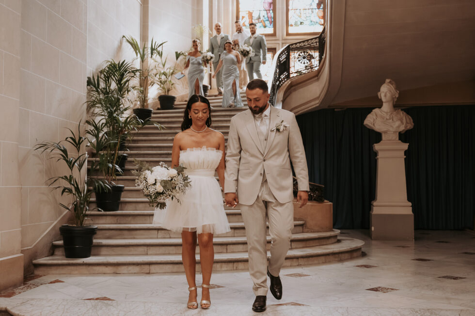 Les mariés descendent le grand escalier de la mairie après la cérémonie civile, la mariée porte une robe courte.