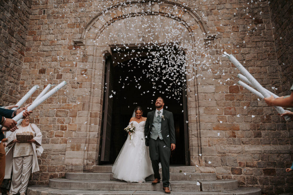 Sortie d'église joyeuse sous une pluie de confettis en forme de cœurs blancs. Le marié porte un costume vert foncé.